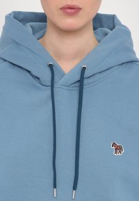Personne portant un sweat à capuche bleu clair avec des cordons bleu foncé et un petit patch zèbre coloré sur la poitrine, lèvres visibles mais yeux hors champ.