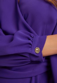 Fioletowa bluzka z marszczonymi rękawami i złotym guzikiem. Materiał ma gładką teksturę i elegancki układ.
