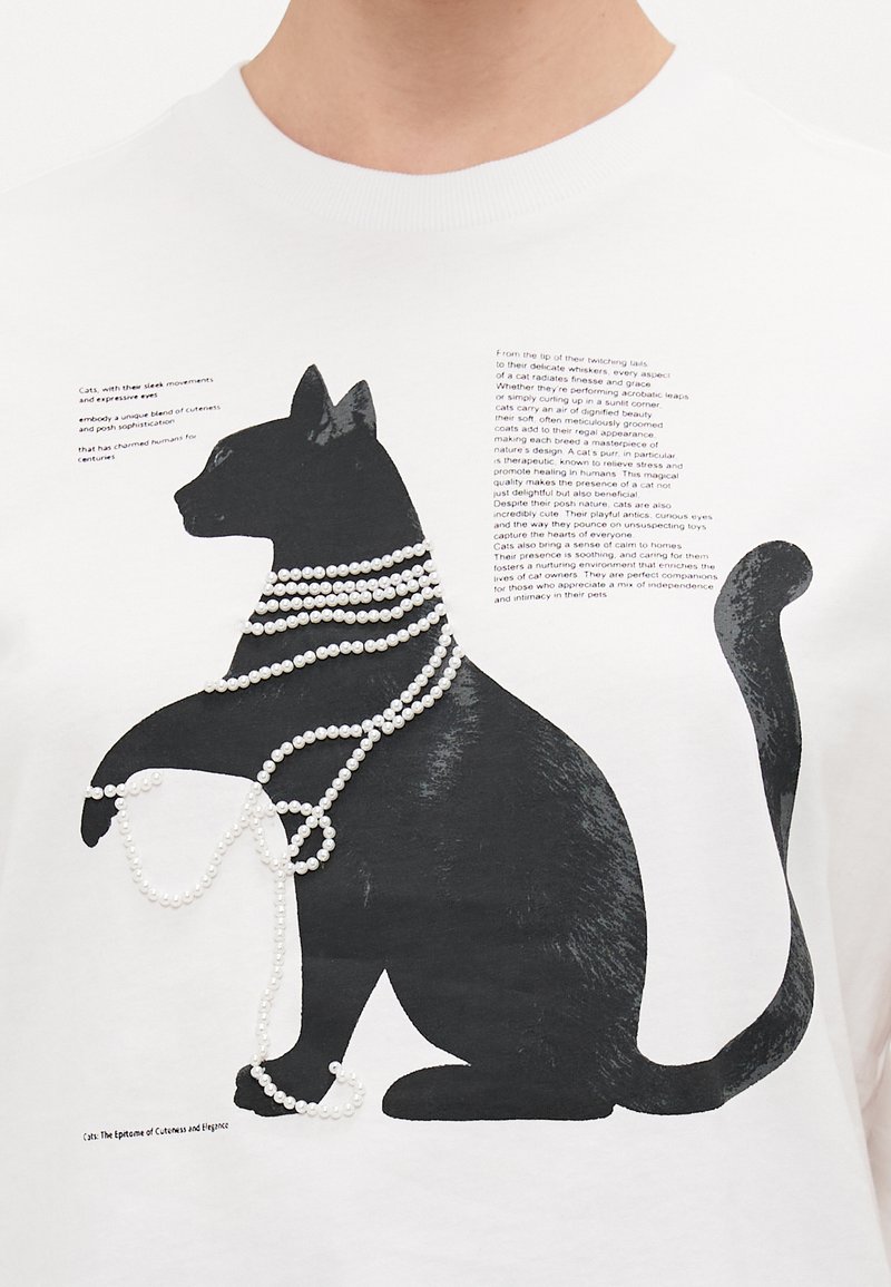 Weißes T-Shirt mit einem großen schwarzen Katzenmotiv, das mit weißen perlenähnlichen Perlen verziert ist, begleitet von schwarzem gedrucktem Text.
