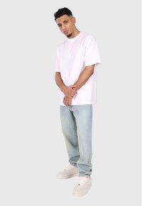 Hvid kortærmet t-shirt, lyseblå denim jeans og hvide sneakers. Modellen står med armene over kors foran en neutral baggrund.