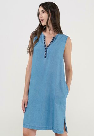 Mujer con vestido sin mangas de mezclilla azul claro con ribete estampado en el escote en V, manos en los bolsillos laterales, mirando hacia su izquierda.