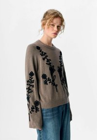Junge Frau mit lockigem Haar, die einen taupefarbenen Pullover mit schwarzer Blumenstickerei und locker sitzenden blauen Jeans trägt, vor einem einfarbigen Hintergrund.