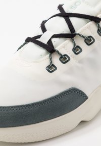 Baskets blanches avec une tige en tissu lisse, des accents en suède gris, des lacets noirs et des œillets verts. Semelle texturée avec un motif subtil.