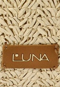 Fondo tejido de paja natural con una etiqueta de cuero marrón que presenta la palabra "LUNA" estampada en oro. Textura en un tono beige claro.