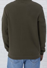 Camisa de manga larga de color verde oliva texturizado con un diseño acanalado y un corte holgado, llevada sobre unos jeans de mezclilla azul claro.