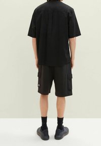 Osoba stojící zády, oblečená v černém tričku s krátkým rukávem, černých cargo šortkách s kapsami, černých ponožkách a černých teniskách.