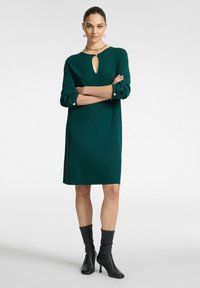 Grün gestricktes Kleid mit dreiviertelärmeln, Schlüsselloch-Ausschnitt und Knopfakzenten. Getragen mit schwarzen kniehohen Stiefeletten mit Absatz.