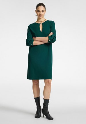 Abito lavorato a maglia verde con maniche a tre quarti, scollatura a chiave e dettagli con bottoni. Indossato con stivali neri al ginocchio con tacco.