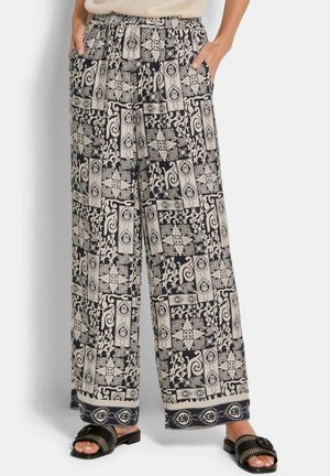 Vrouw draagt losse zwarte en beige broek met patroon, beige top ingestopt, handen in de zakken en zwarte slippers met gesp.