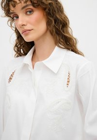 Weißes Hemd mit Knöpfen und Perlenverzierungen sowie gestickten Paisley-Mustern. Strukturierter Stoff und Spitzenausschnitte vervollständigen das Design.