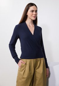Marineblå omslagsgenser laget av myk stoff, med v-hals og lange ermer, parret med plisserte beige bukser.