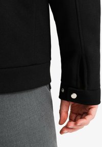 Veste noire texturée avec une taille cintrée, dotée d'un design de poignet à boutons-pression. Pantalons gris visibles au bord inférieur.