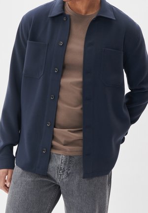 Hombre con chaqueta azul oscuro desabrochada sobre camiseta marrón y vaqueros grises, de pie con una mano detrás de la espalda.