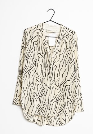 Lekka, kremowa bluzka z czarnymi, falistymi paskami. Ma dekolt w kształcie litery V, długie rękawy i zaokrąglony dół. Wisi na czarnym haczyku.