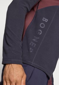 Maglia sportiva a maniche lunghe di colore blu navy scuro con accenti bordeaux. Presenta un tessuto testurizzato, dettagli di cucitura visibili e un logo stampato sul lato.