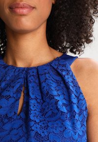 Detailní záběr ženy v bezrukávovém modrém krajkovém topu s květinovými vzory a malým výstřihem ve tvaru klíče.