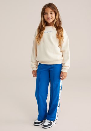 Creme sweatshirt med teksten "Young and Free", parret med blå træningsbukser med hvide sidelister og sorte stjerner. Sorte og hvide sneakers.