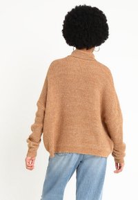 Brauner Strickpullover mit hohem Kragen und weiten Ärmeln, kombiniert mit hellblauen Jeans. Der Stoff hat eine strukturierte Oberfläche.