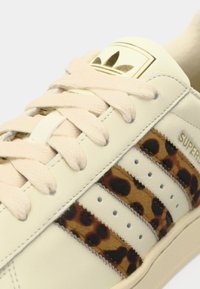 Sneakers with a cream leather upper, light pink laces, and brown leopard print stripes. Features three perforated stripes and gold branding.