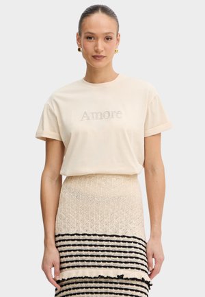 Donna con i capelli raccolti in uno chignon, che indossa orecchini a cerchio dorati, una maglietta beige a maniche corte con la scritta "Amore" e una gonna a righe beige e nere testurizzata.