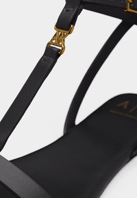 Sangle de sandale en cuir noir avec boucle en métal doré ornée d'un détail de logo stylisé "A|X".