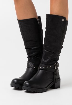 Bottes noires mi-mollet avec talons blocs bas, fermetures éclair latérales, sangles cloutées et boucles décoratives portées sur des jambes nues sur un fond blanc.