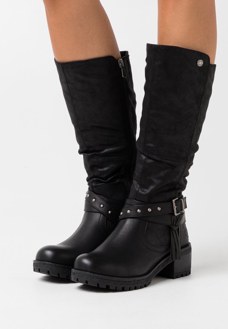 Bottes noires mi-mollet avec talons blocs bas, fermetures éclair latérales, sangles cloutées et boucles décoratives portées sur des jambes nues sur un fond blanc.