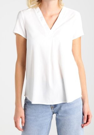 Vrouw draagt een witte V-hals blouse met korte mouwen, gecombineerd met lichtblauwe spijkerbroek, getoond van schouders tot heupen.