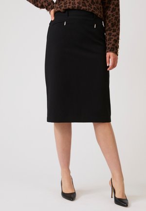 Femme portant une jupe crayon noire longueur genou avec poches zippées, un haut marron imprimé léopard, et des chaussures à talons noirs.