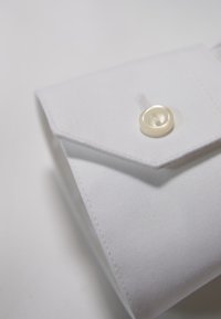 Polsino di camicia bianca in cotone con forma appuntita, un solo bottone in madreperla e dettagli di cuciture visibili. Tessuto liscio.