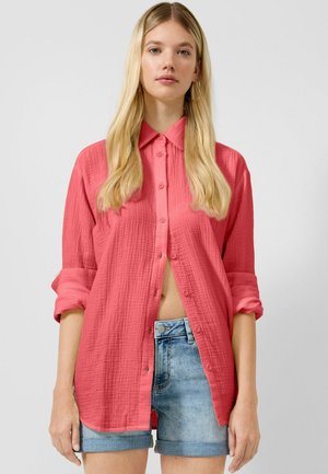 Jeune femme portant une chemise ample corail à boutons avec les manches retroussées et un short en jean bleu clair sur un fond uni.