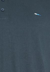 Polo bleu sarcelle foncé avec trois boutons et un petit rectangle brodé en blanc et bleu sur le côté gauche de la poitrine.