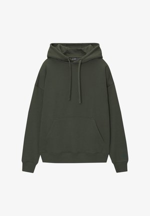 Persona con sudadera con capucha verde oscuro, jeans cargo holgados de color azul claro y zapatillas marrones oscuras, de pie con una mano en el bolsillo.