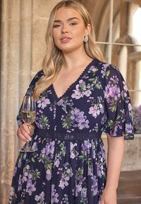 Kukkakuosisia mekko tummansinisellä taustalla, jossa on violetteja ja vihreitä yksityiskohtia. Mekossa on V-kaula-aukko, pitsireunus ja ilmavat lyhyet hihat, ja se on yhdistetty lasilliseen samppanjaa.