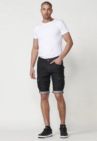 Schwarze Denim-Cargo-Shorts mit umgeschlagenen Säumen, ausgestattet mit Fronttaschen und einem Knopfverschluss. Tragen mit einem eng anliegenden weißen T-Shirt und schwarzen Schuhen.