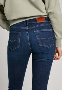 Jeans de denim de color azul oscuro con un diseño ajustado, que cuentan con dos bolsillos traseros, costuras en contraste y un parche de cuero marrón de la marca en la cintura.