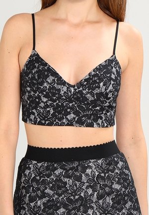 Kobieta ubrana w czarny koronkowy krótki top na cienkich ramiączkach i dopasowaną spódnicę z wysokim stanem, odsłaniającą brzuch oraz detale z kwiatowym wzorem.