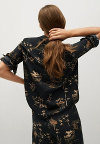 Camisa negra con estampado floral y paisajístico dorado, con un corte relajado, puños enrollados y una textura suave.