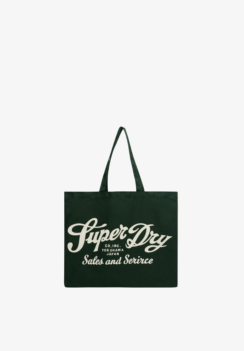 Sac cabas vert foncé avec texte beige manuscrit « Super Dry Co. Inc. Yokohama Japan Sales and Service » et deux anses d’épaule.
