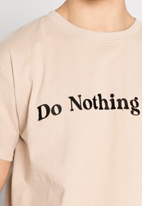 Beige tričko s černě vyšitým textem "Nedělej nic", nošené osobou, zobrazené od horní části hrudníku a části krku.