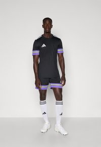 Tenue de football noire avec des manches courtes et un short, ornée de rayures violettes et vertes. Chaussettes blanches et chaussures avec des accents noirs.