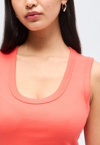 Γυναίκα που φοράει κοραλί αμάνικη ριγέ μπλούζα και ασημένια σκουλαρίκια σε σχήμα καρδιάς, με μακριά σκούρα μαλλιά και φυσικό μακιγιάζ.