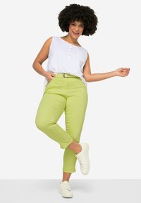 Grüne Baumwollmischungshose mit verkürztem Schnitt, kombiniert mit einem weißen ärmellosen Oberteil mit Knopfdetails und einer Brusttasche. Weiße Sneakers.