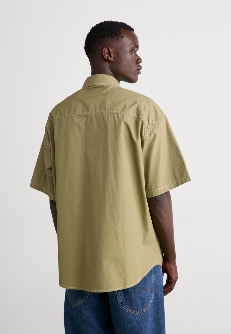 Camisa de manga corta color verde oliva con cuello, que presenta un corte relajado y detalles de costura visibles. Combinada con jeans de mezclilla azul holgados.