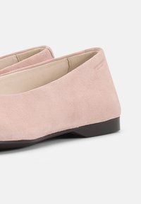 Ζευγάρι παιδερών φλατ παπουτσιών από ανοιχτό ροζ σουέντ με χαμηλά μαύρα τακούνια, μπεζ εσωτερική επένδυση και διακριτικό ανάγλυφο λογότυπο στο εξωτερικό μέρος.