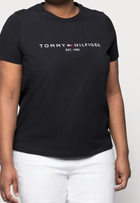 Sort bomulds T-shirt med rund hals, med hvid "TOMMY HILFIGER" logo og "EST. 1985" tekst i midten, båret med hvide bukser.