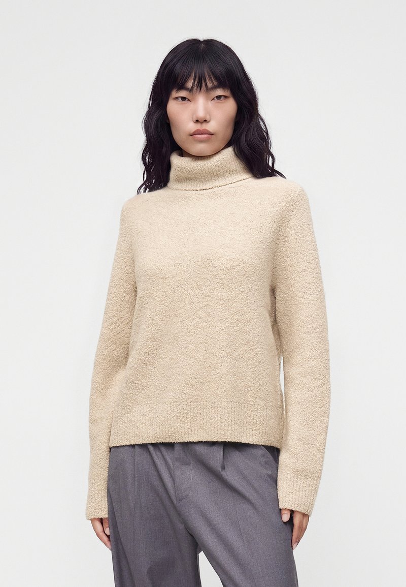 Femme aux longs cheveux noirs portant un pull à col roulé beige et un pantalon gris, debout devant un fond blanc uni.