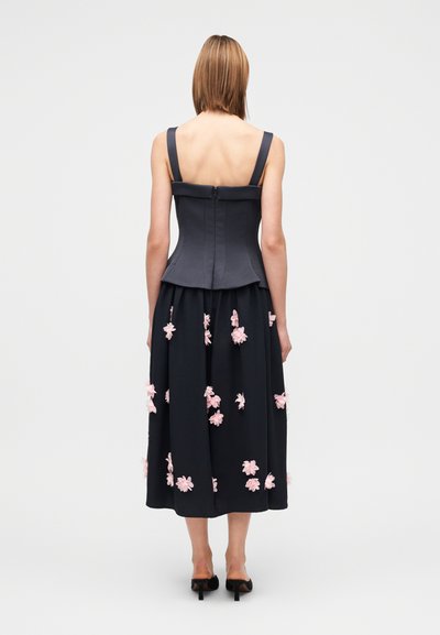 Σκούρο γκρι κορσέ τοπ με χοντρές τιράντες, σε συνδυασμό με μια ρέουσα μαύρη φούστα διακοσμημένη με ροζ φλοράλ εφαρμογές. Ορατή λεπτομέρεια φερμουάρ στην πλάτη.