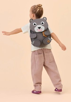 Child with hair in bun wearing a gray bear-shaped backpack, extending arms, dressed in white shirt, pink pants, and purple shoes on beige background.