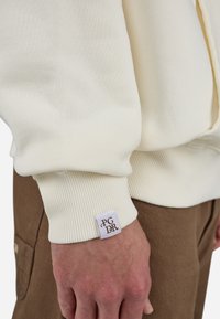 Cream-colored sweatshirt featuring ribbed cuffs and a woven label with "PGDR" on the left wrist. Soft texture, relaxed fit.